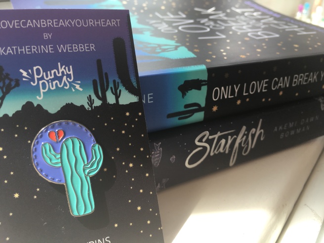 The spines of the books Only Love Can Break Your Heart and Starfish. An enamel pin with a cactus and a broken heart is in the foreground.